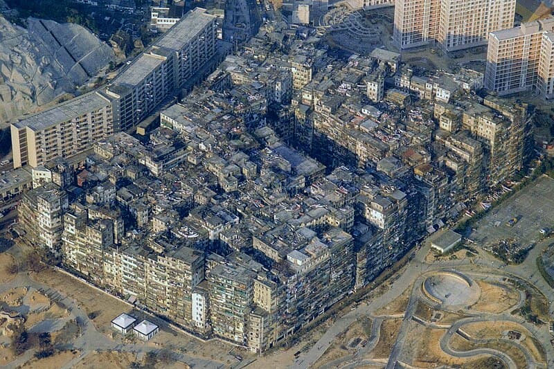 Aerial photograph of Kowloon Walled City in 1989: a single massive rectangular block of gray buildings fused together, with Kai Tak Airport's runway and planes visible in the background and ordinary Hong Kong suburbs surrounding it on all sides.
