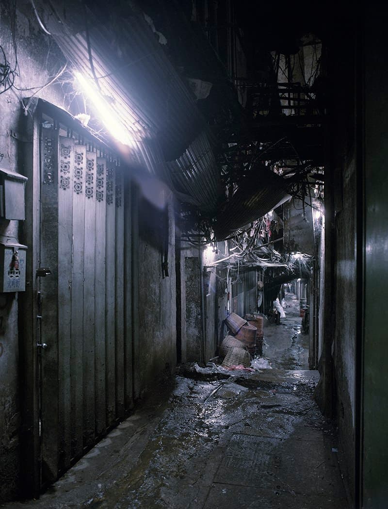 An interior alley inside Kowloon Walled City in 1993: a narrow, low-ceilinged passage lit only by scattered fluorescent tubes, with exposed pipes and wiring running across every wall and the ceiling.